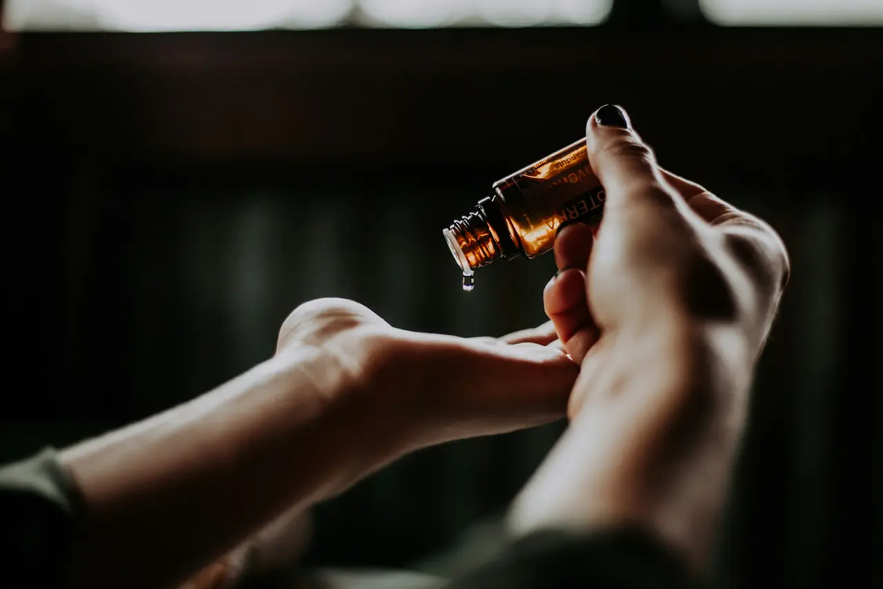 Drops of essential oil fall from an amber bottle into a waiting palm, in warm low light.