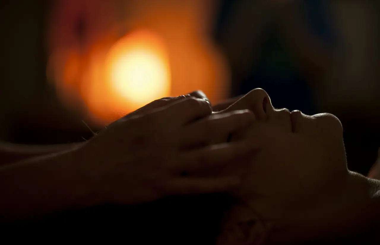 Hands rest lightly on a person's face during a massage, lit by the glow of a warm lamp.