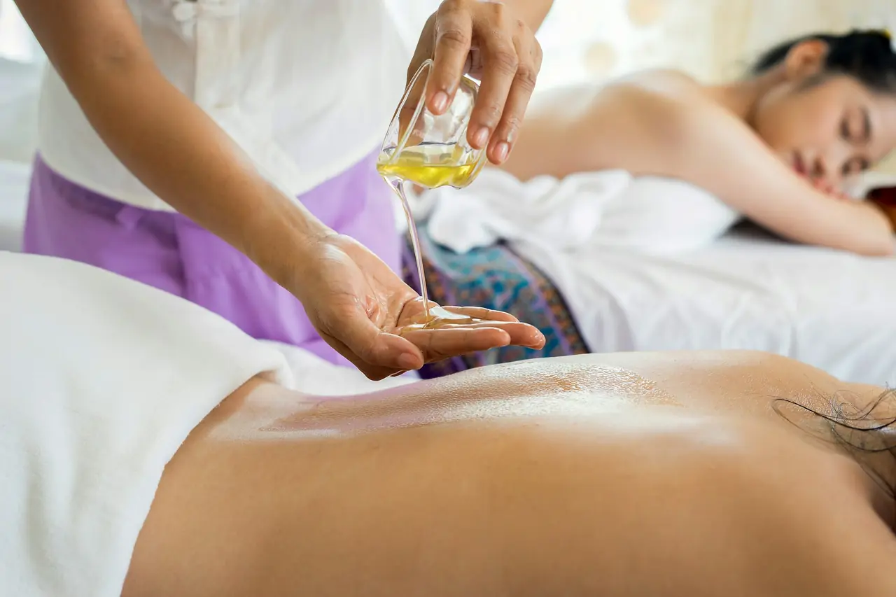 A therapist pours warm massage oil into her palm beside a woman resting face down on a white treatment bed.